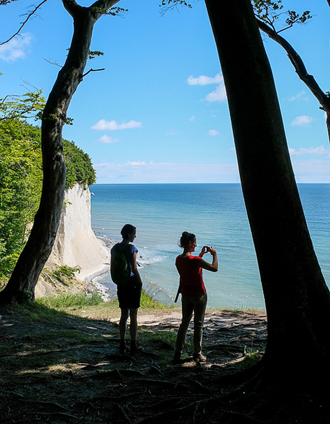 Zwei Personen stehen am Rande einer Klippe mit Blick auf das Meer. Die Landschaft ist von Bäumen umgeben, und einer der Personen macht ein Foto. Das Wetter ist klar mit blauen Himmel und wenigen Wolken.
